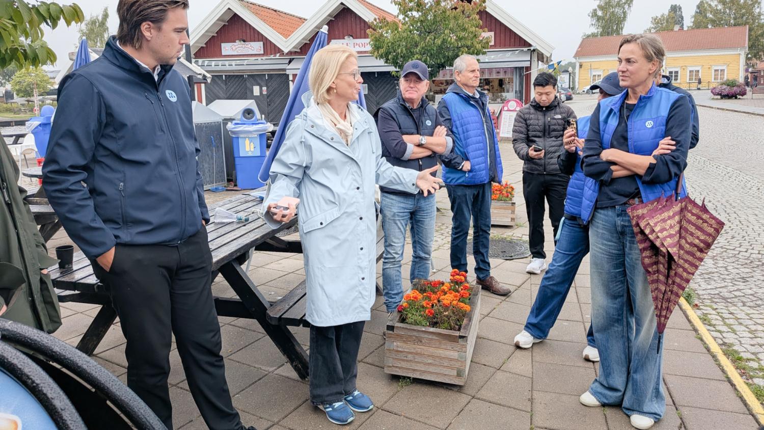 En grupp människor klädda i jackor och jeans står och pratar på en stenlagd trottoar bredvid blomlådor och trähus.
