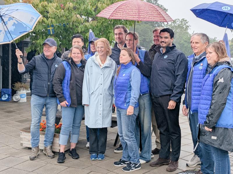 En grupp människor klädda i blå västar och jackor står tillsammans utomhus en regnig dag; några håller i paraplyer och poserar för ett foto.