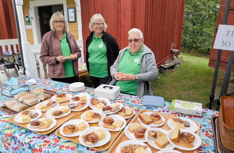 Tre personer står bakom ett bord med olika bakverk och brödbullar på tallrikar vid en matbod utomhus, med röda och gula byggnader och grönska i bakgrunden.