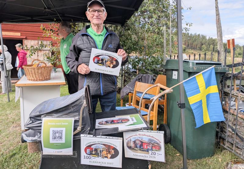 En äldre man vid ett utomhusstånd säljer kalendrar med svenskt tema och har en svensk flagga uppsatt. På bordet syns en QR-kod för mobilbetalning.