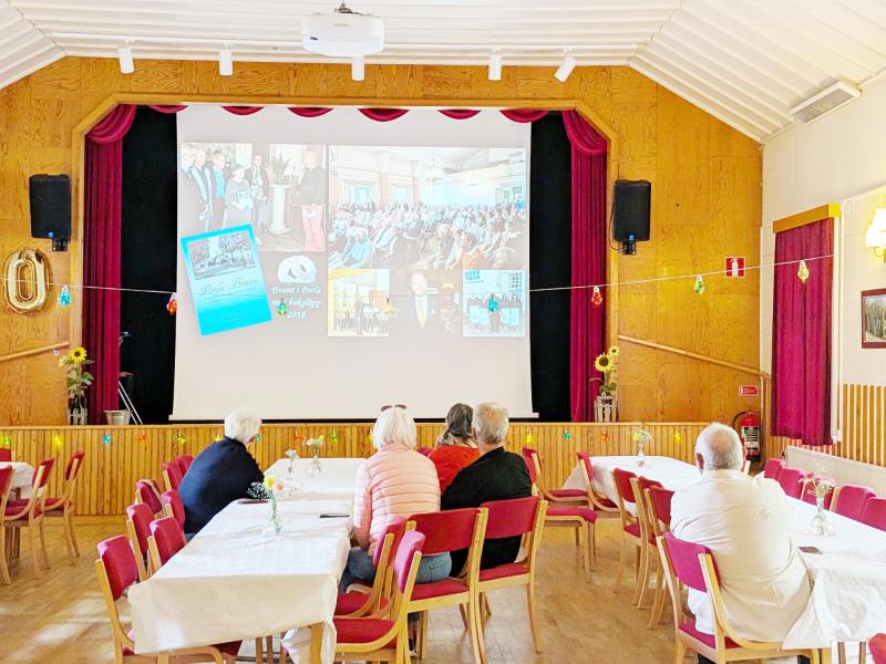 Fem personer sitter vid bord i en sal och tittar på ett bildspel som projiceras på en stor skärm längst fram i rummet. Salen har träväggar, röda stolar och festliga dekorationer.