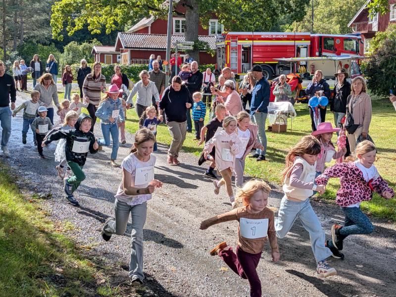 En grupp barn med numrerade startnummer springer ett lopp på en grusväg, medan vuxna och andra barn tittar på; i bakgrunden syns en brandbil och röda byggnader.