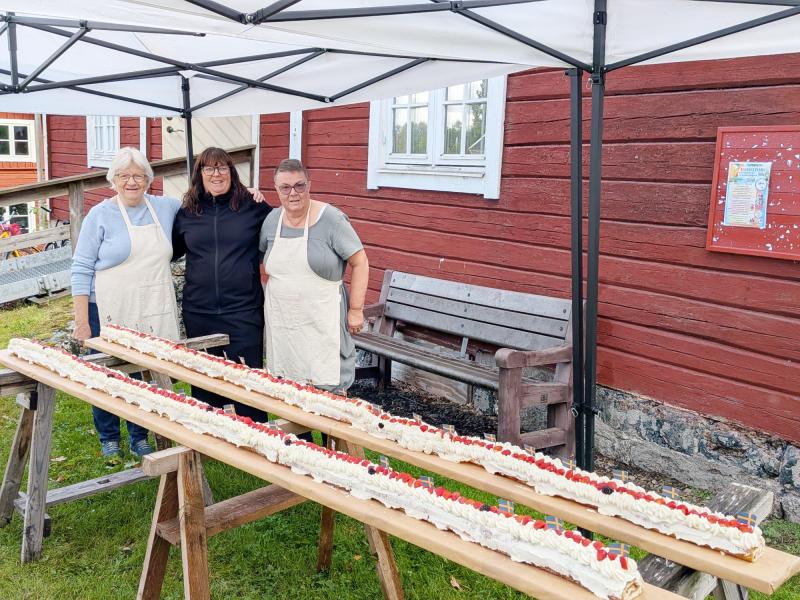 Tre kvinnor står under ett tält bredvid en lång tårta med grädde och bär som prydnad, placerad på träbrädor utanför en röd träbyggnad.