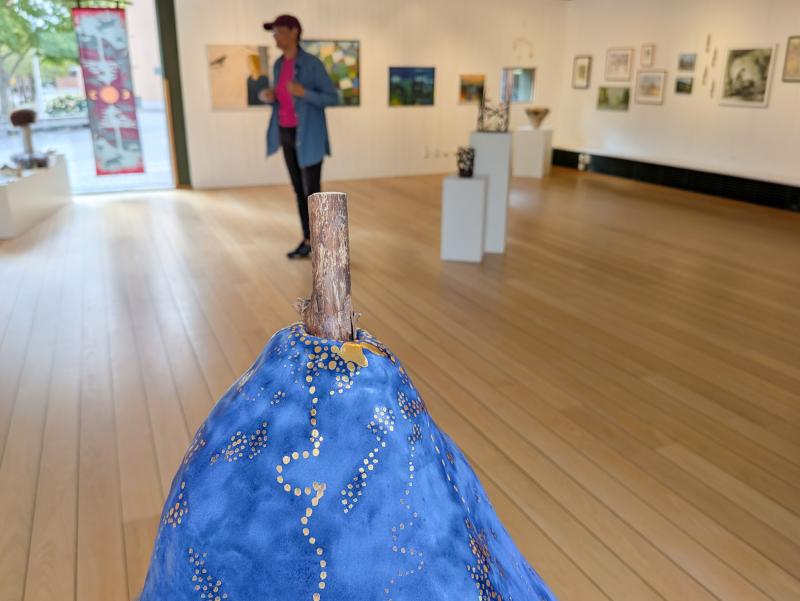 En blå och guldfärgad päronformad skulptur i förgrunden i ett konstgalleri, med en person som står och betraktar konstverken på väggarna i bakgrunden.