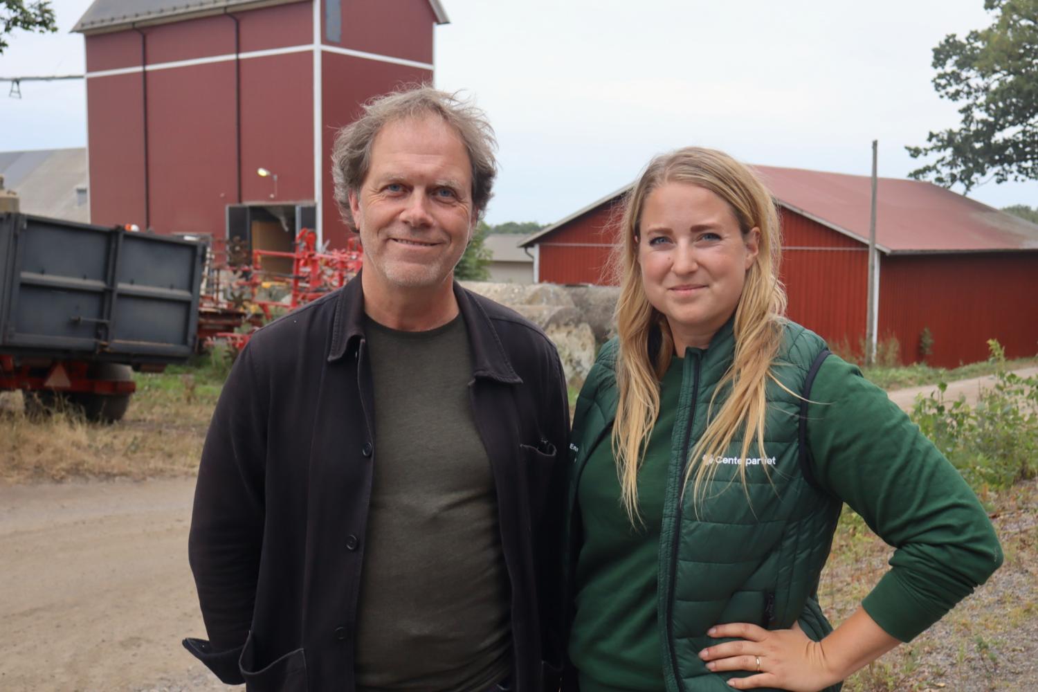 Två personer står utomhus på en grusväg framför röda lantbruksbyggnader och poserar för kameran. Det verkar vara en lantlig miljö.