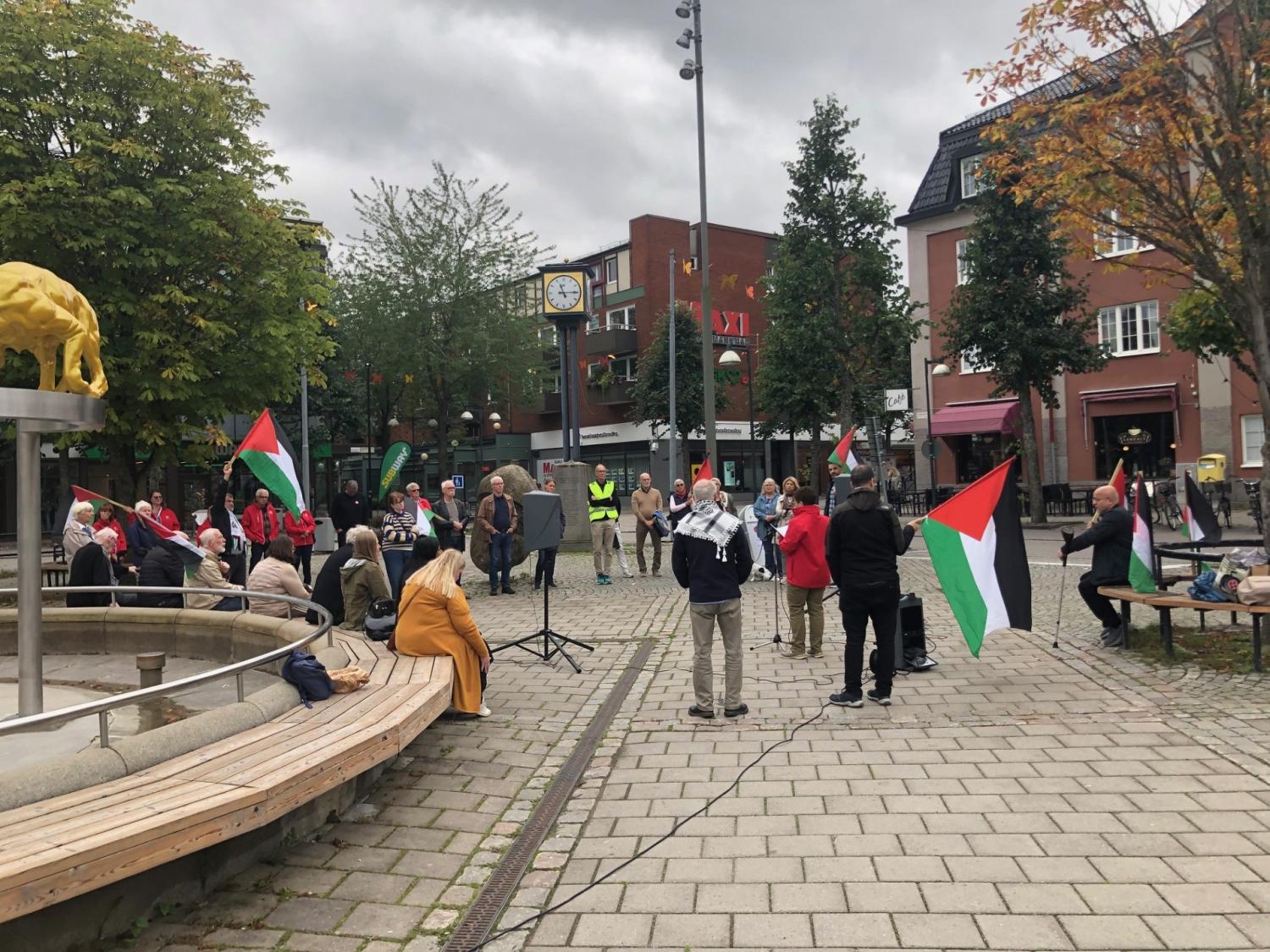 En liten grupp människor samlas på ett torg med palestinska flaggor vid en fontän, några sittande och andra stående, omgivna av träd och byggnader.
