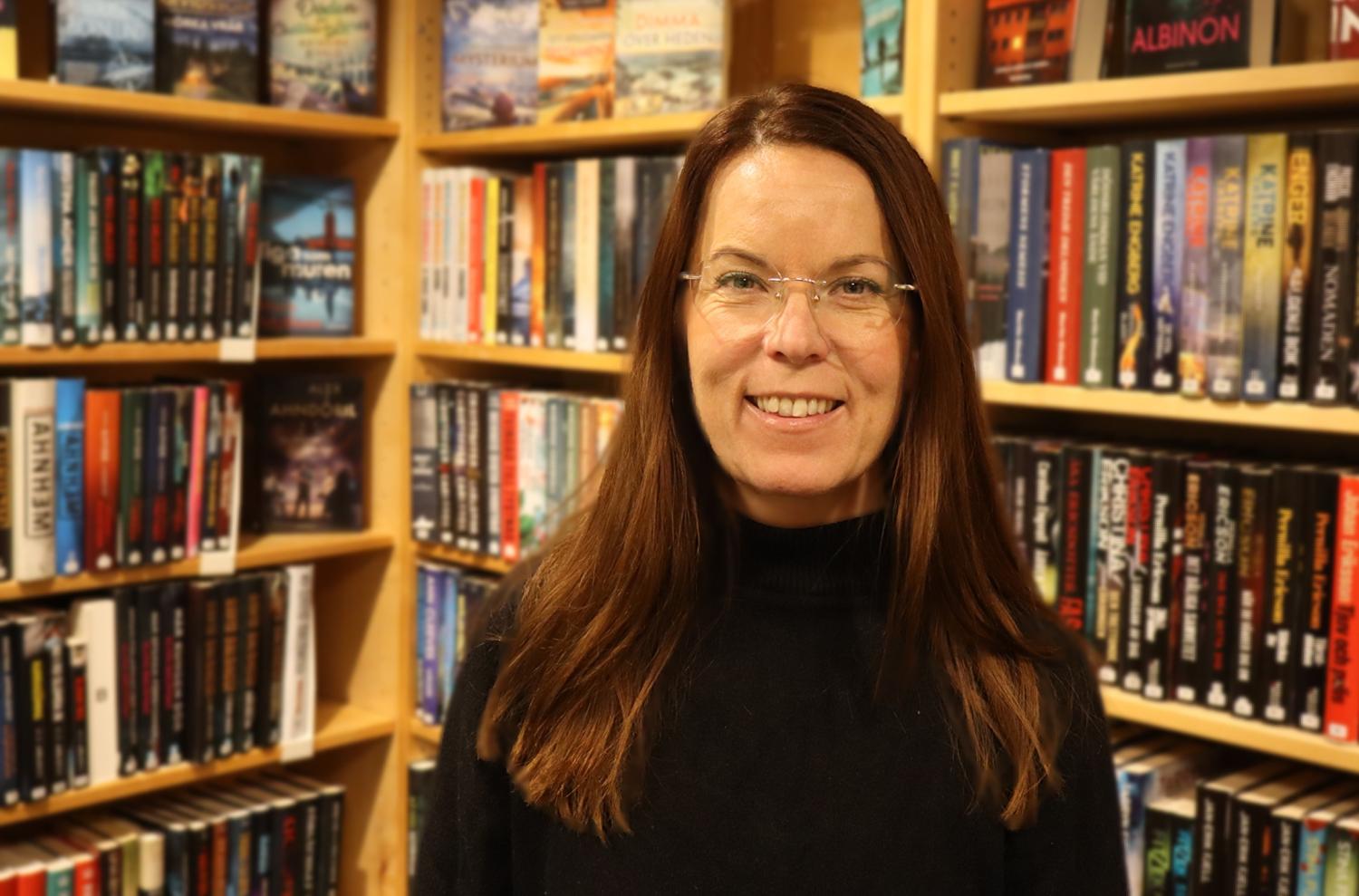 En kvinna med långt brunt hår och glasögon står leende framför bokhyllor fyllda med olika böcker på ett bibliotek eller i en bokhandel.