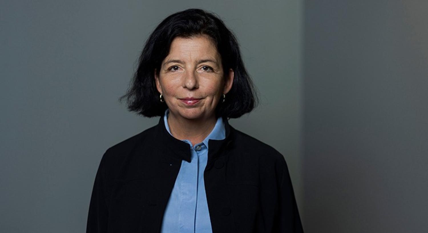 En kvinna med axellångt svart hår, klädd i en blå skjorta med krage och en svart jacka, som står framför en enfärgad grå bakgrund.