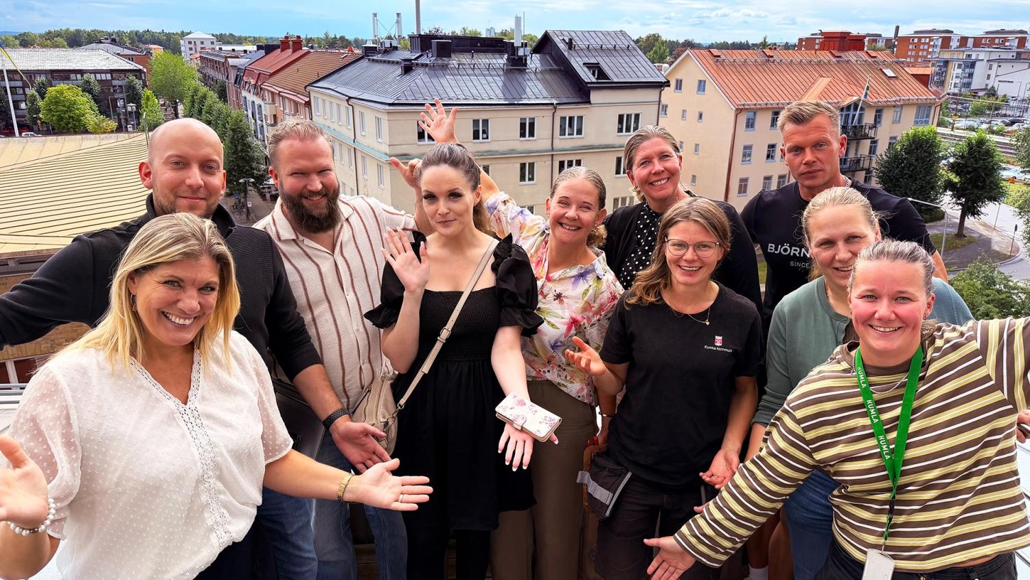 Tio personer står på en balkong och ler mot kameran, medan byggnader, träd och en delvis molnig himmel syns i bakgrunden.