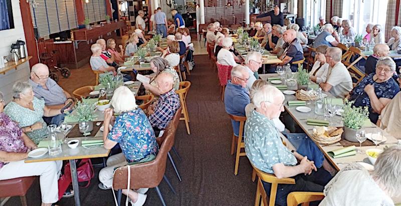 En stor grupp äldre sitter vid långbord i en restaurang och pratar och äter tillsammans.