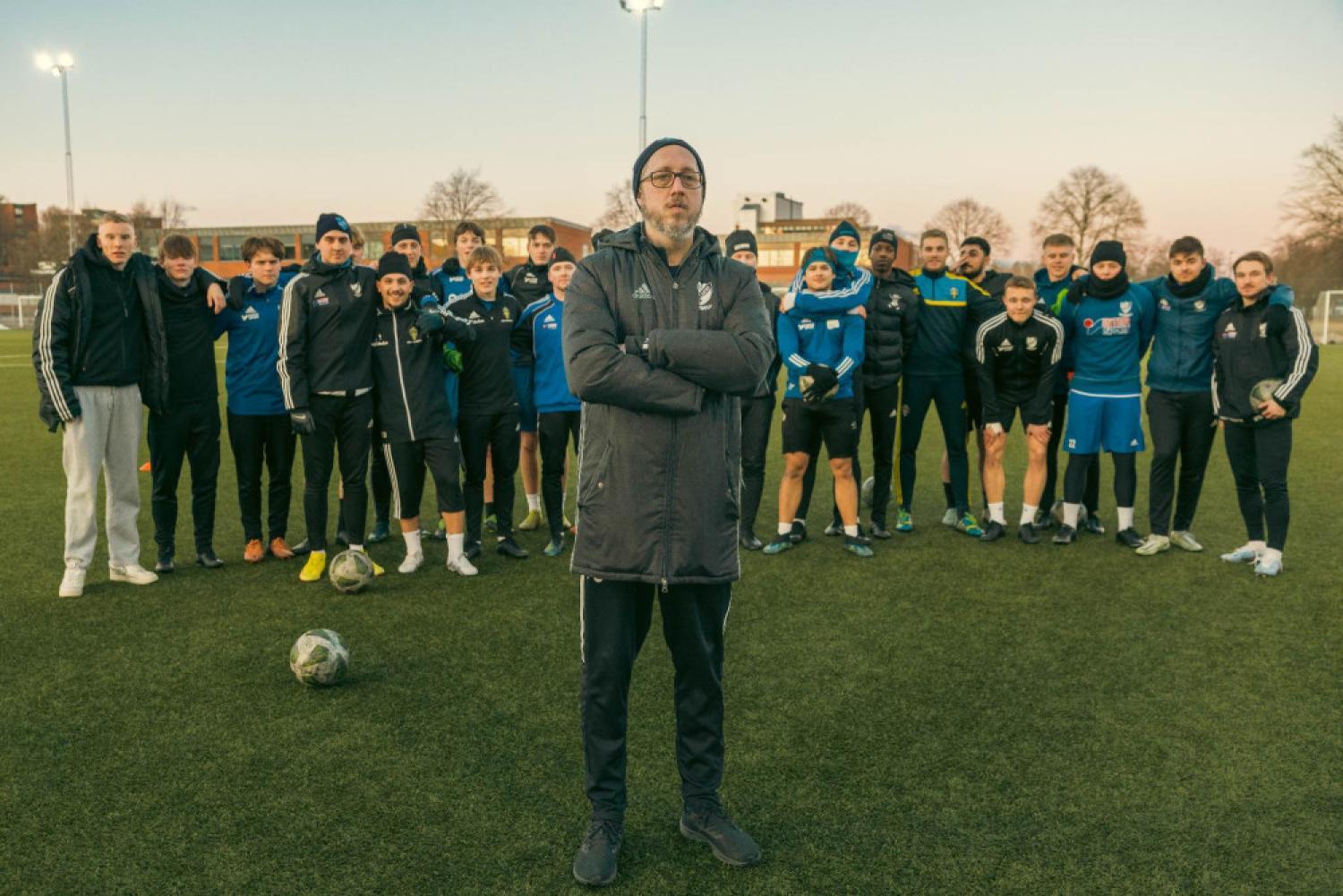 En fotbollstränare står framför en grupp spelare på en utomhusplan. Tränaren är klädd i en mörk rock och en mössa, och spelarna är klädda i atletisk utrustning, några med vinterkläder.