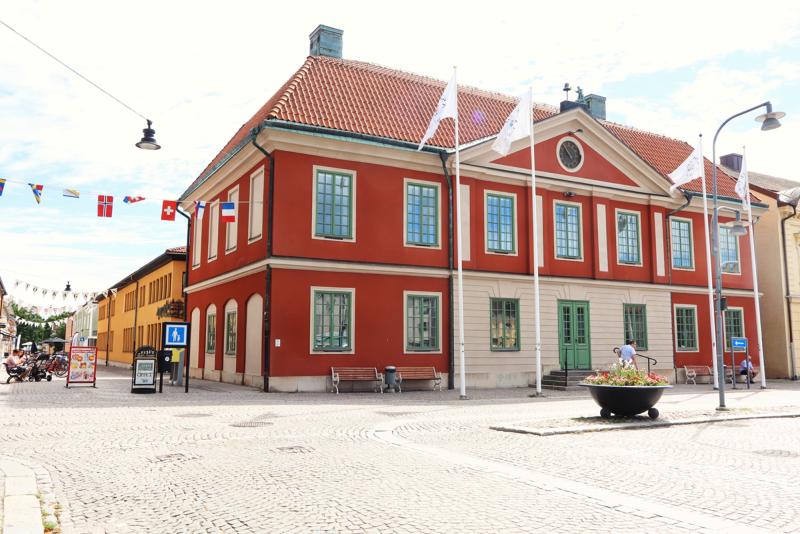 En röd tvåvåningsbyggnad med gröna dörrar och fönster med vita lister står på en kullerstensgata, med flaggor som hänger ovanför och bänkar samt en blomlåda framför.