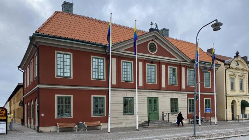 En röd tvåvåningsbyggnad med gröna fönster, svenska flaggor, en klocka ovanför entrén och en person som går på en kullerstensgata framför.