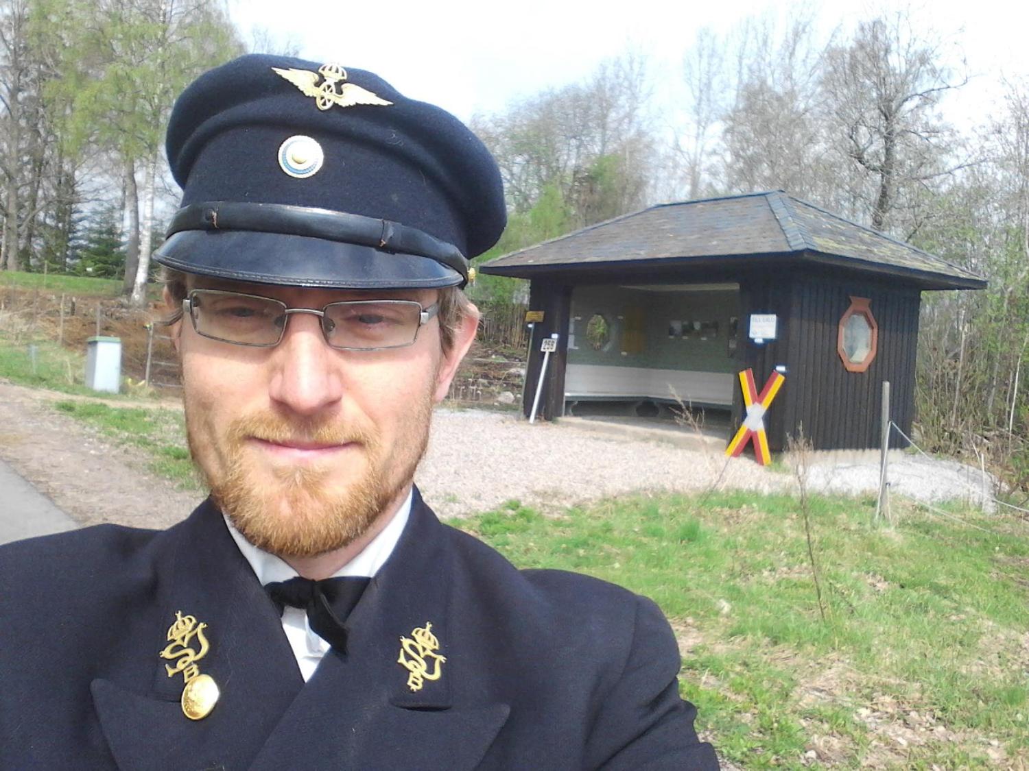 En man i mörk uniform och keps tar en selfie utomhus nära en liten svart byggnad med ett glasfönster och en färgglad stolpe. Träd och gräs syns i bakgrunden.