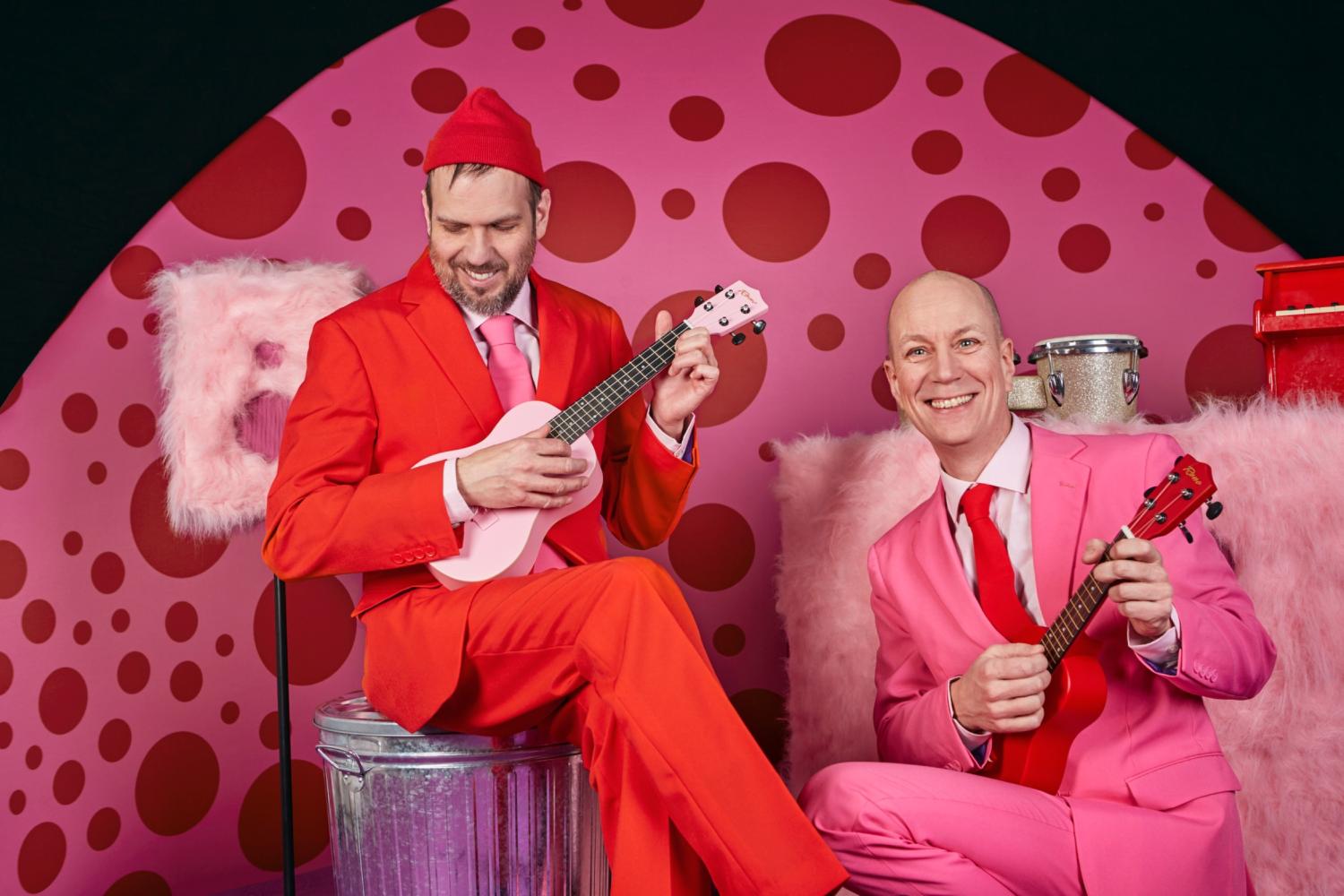 Två män i ljusröda och rosa kostymer spelar ukuleles på en rosa prickig bakgrund, sittande på rosa pälsstolar med musikinstrument i närheten.