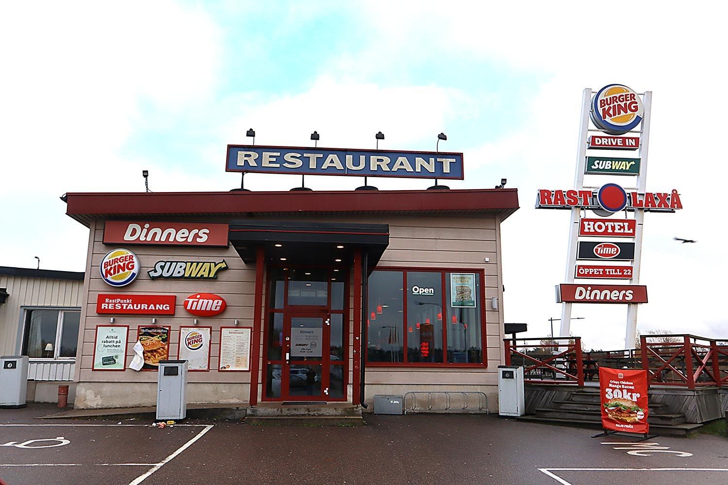 En restaurangbyggnad vid en väg med skyltar för Burger King, Subway, Dinners och Time, och en stor skylt med samma varumärken och hotellinformation.
