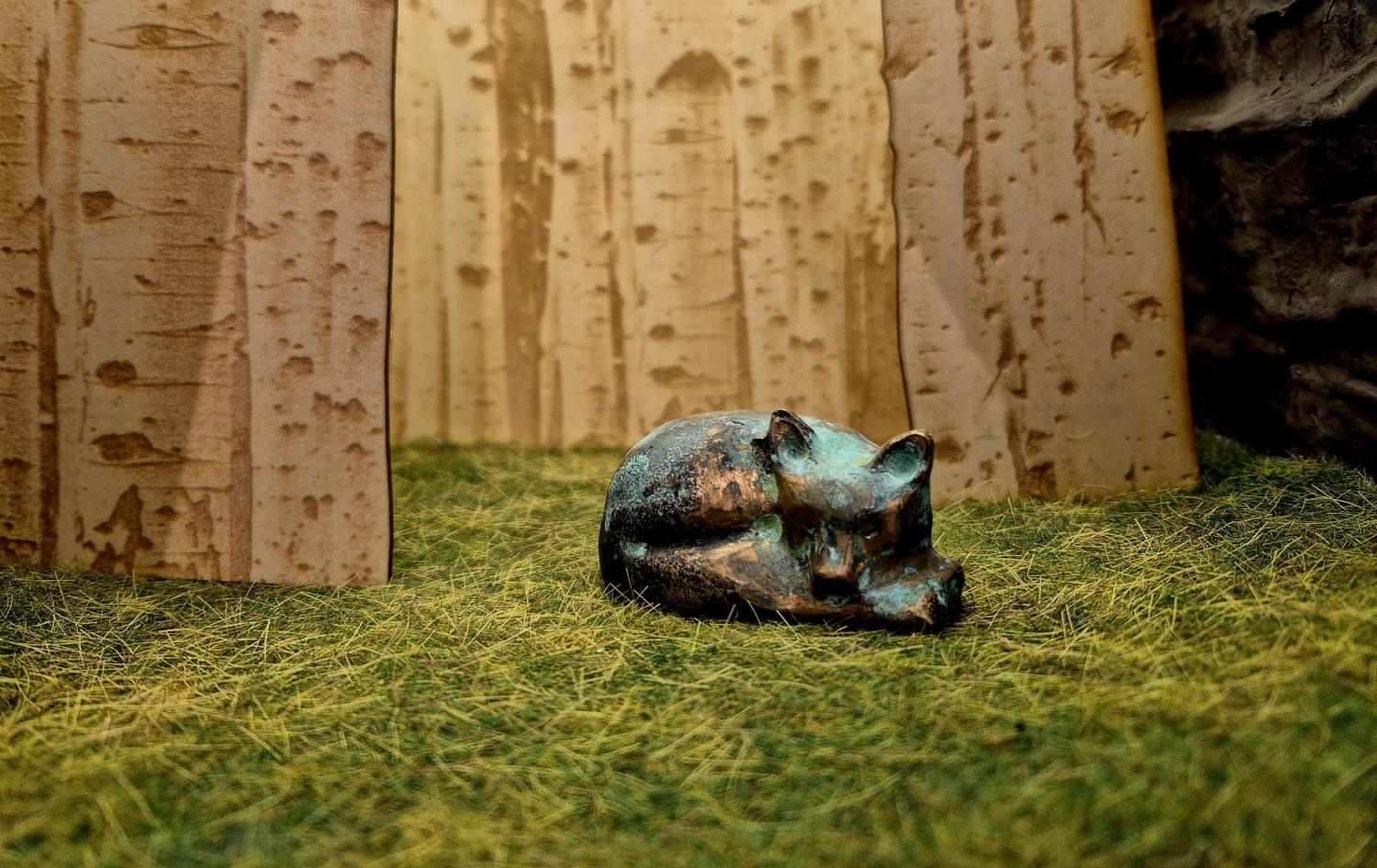 En liten skulptur av en vilande räv sitter på grönt texturerat gräs med en björk i bakgrunden.