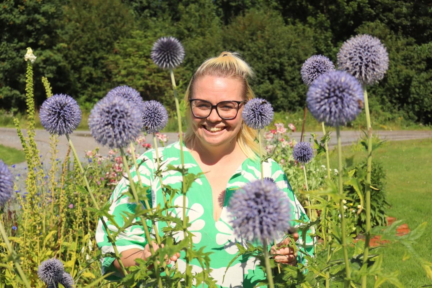 En person med glasögon och en grön blommig skjorta står och ler bland höga, lila alliumblommor i en solig trädgård.