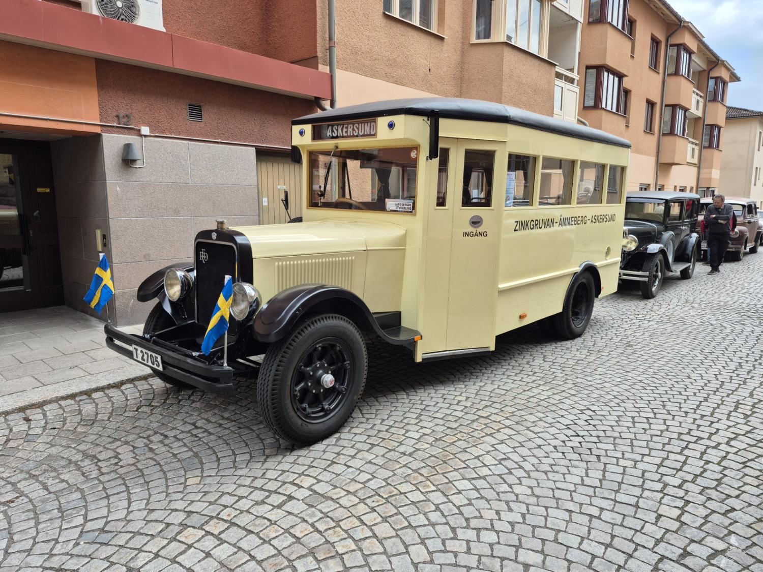 En krämfärgad vintagebuss med svenska flaggor parkerad på en kullerstensgata framför byggnader, med flera klassiska bilar uppradade bakom den.