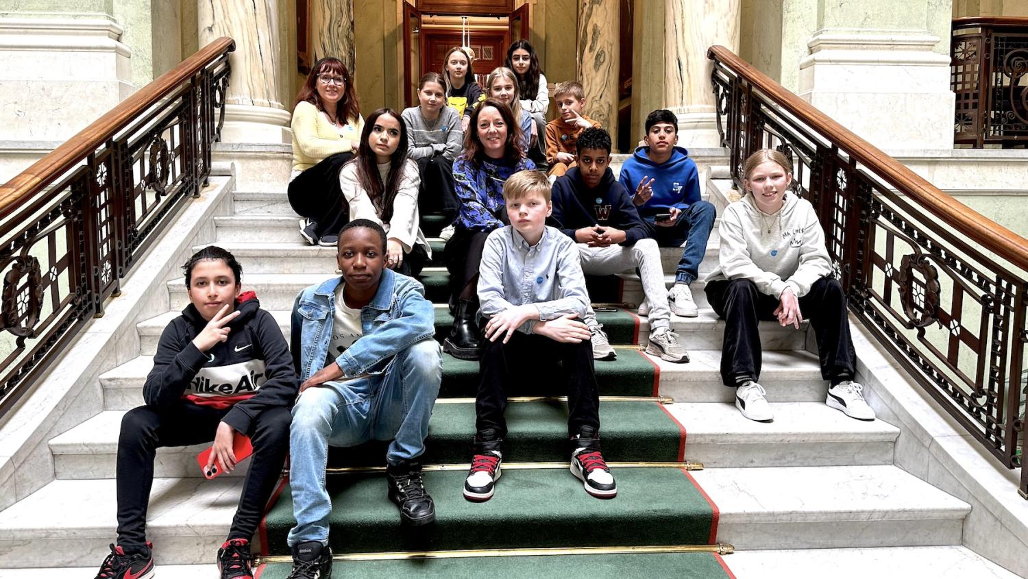 En grupp olika barn som sitter på en trappa med sin lärare bakom dem i en historisk byggnad.