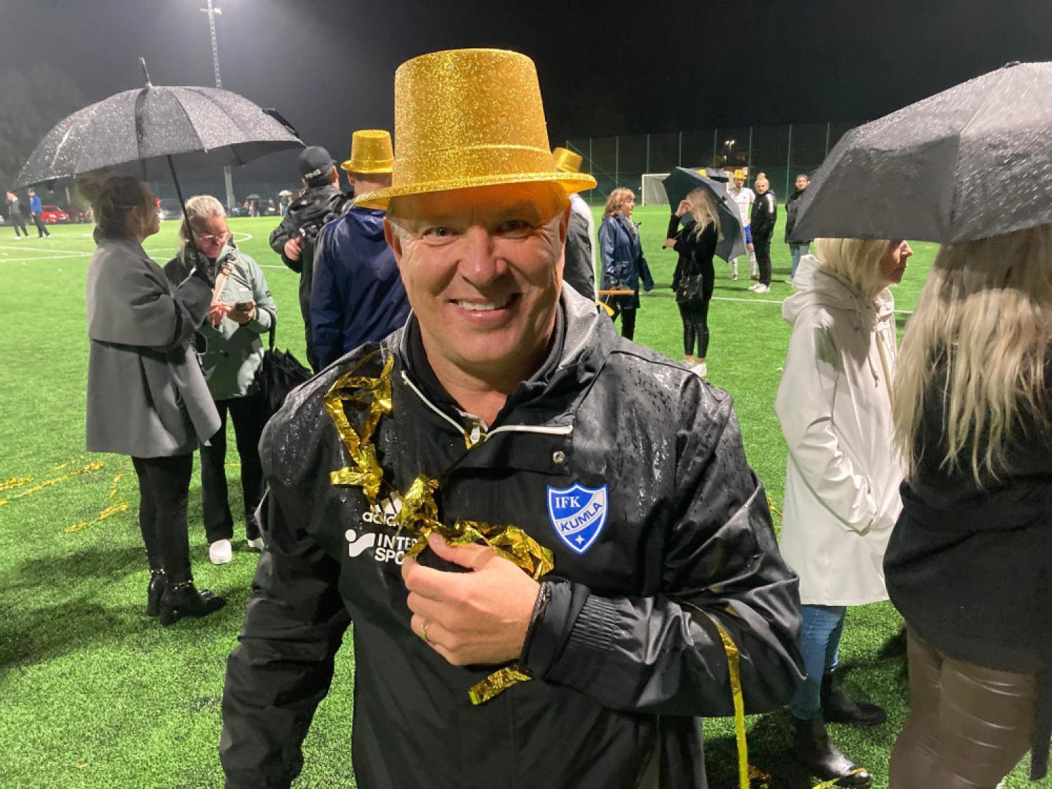 En man med en guldglittrande hatt och en liten pokal står på en våt fotbollsplan med andra människor och paraplyer i bakgrunden.