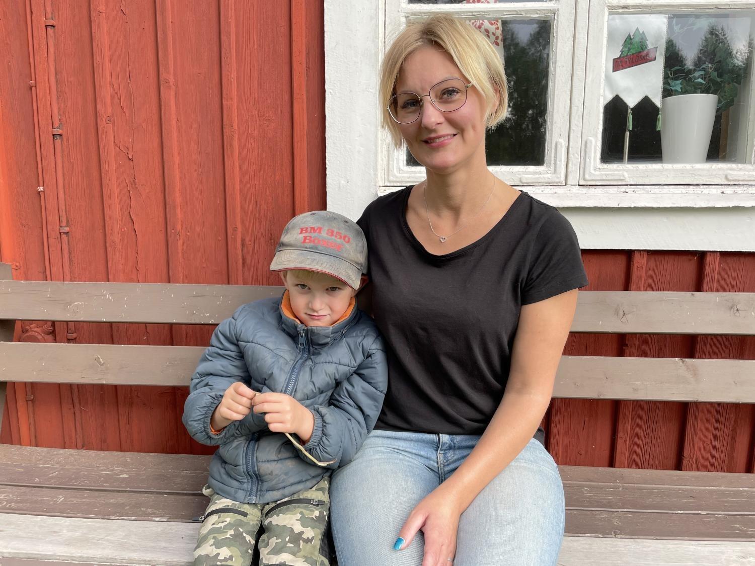 En kvinna med glasögon och en svart tröja sitter på en bänk bredvid en liten pojke med keps och kamouflagebyxor, framför en röd träbyggnad med ett fönster med vit ram.