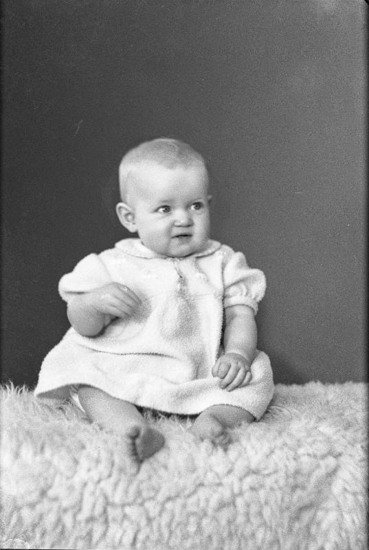 Svartvitt foto av en bebis i en ljus klänning som sitter på en mönstrad filt och tittar lite åt sidan.
