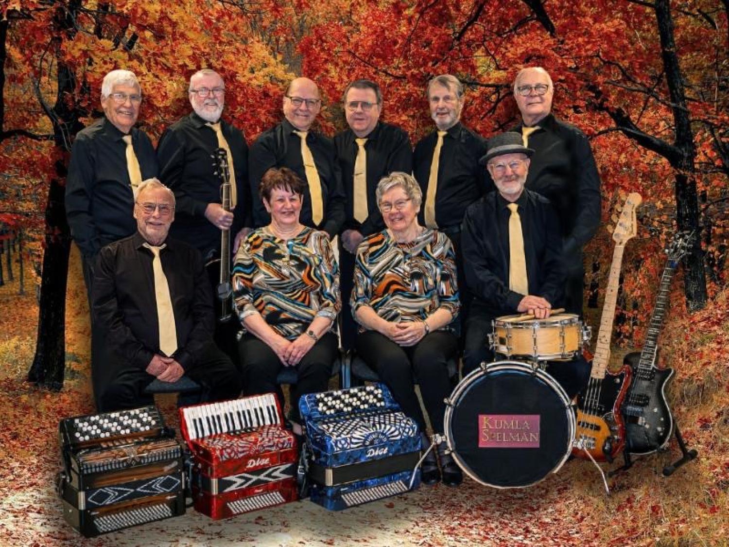 En grupp på tio vuxna i svarta kläder med gula slipsar poserar inomhus med musikinstrument, bland annat dragspel, gitarrer, en klarinett och trummor, mot en höstskogsbakgrund.