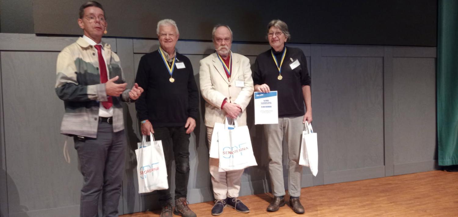 Fyra personer står på en scen; tre bär medaljer och håller i presentpåsar, medan den fjärde, i mönstrad jacka, står bredvid och talar eller presenterar.