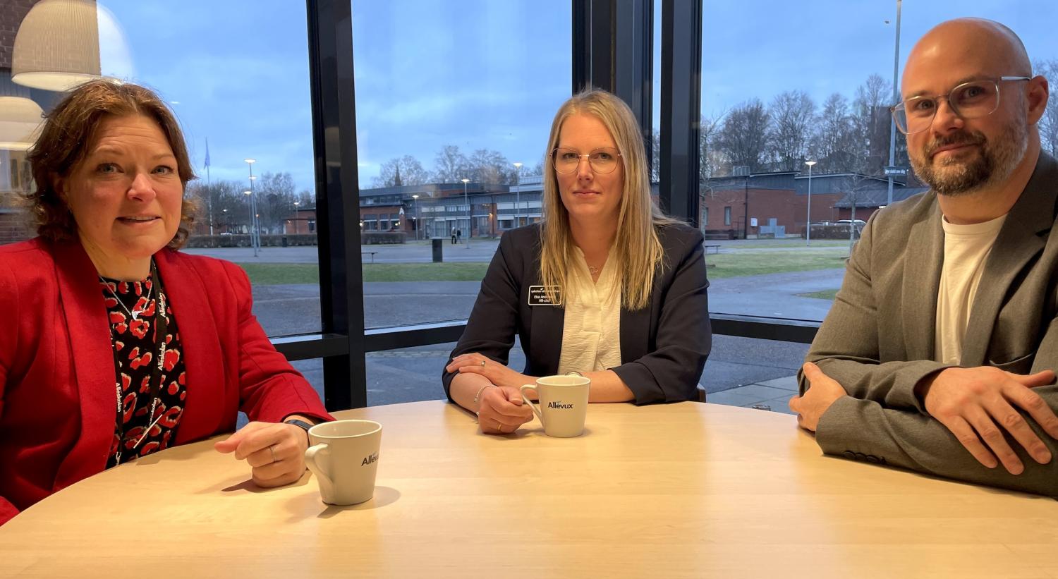 Tre personer sitter vid ett runt bord med kaffekoppar och pratar i ett rum med stora fönster med utsikt över en uteplats.