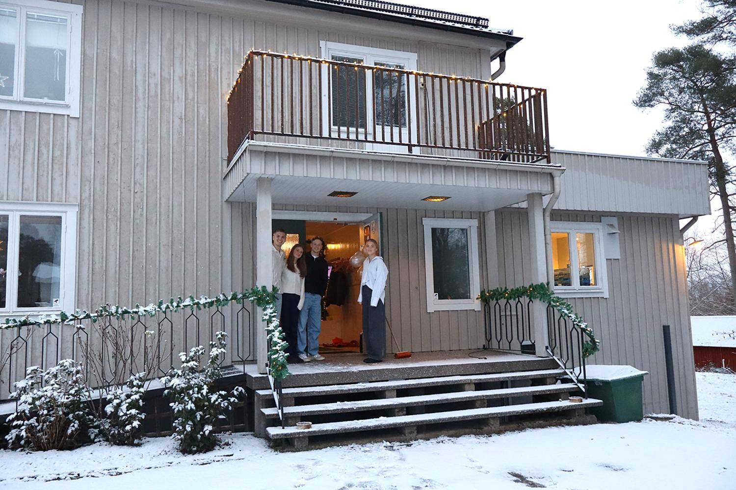 Tre personer står på verandan till ett tvåvånings trähus med snö som täcker marken och trappstegen.