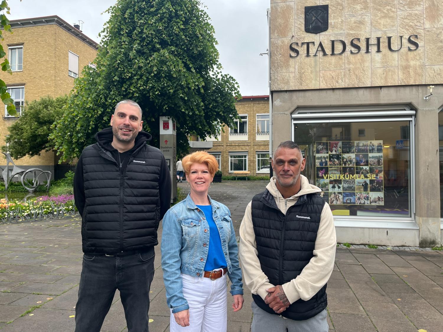 Tre personer står framför en byggnad med en skylt där det står ”STADSHUS”. Gruppen består av två män och en kvinna, alla klädda i vardagsjackor.