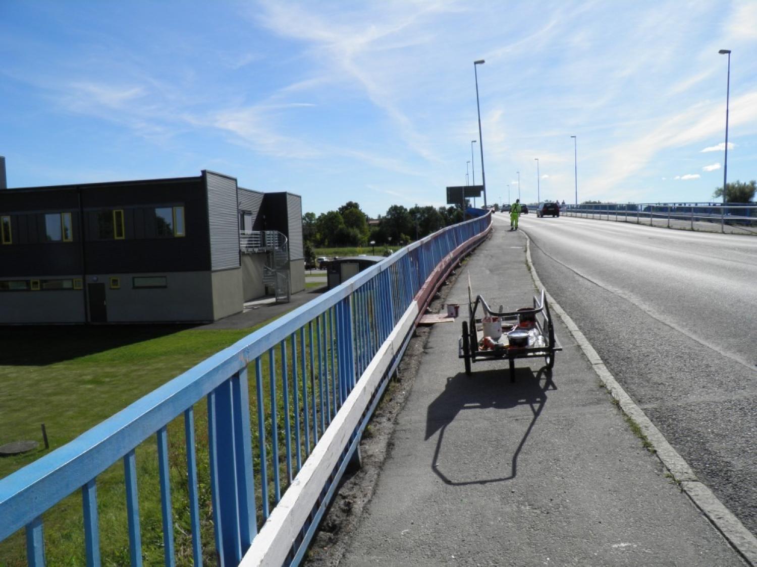 En arbetsvagn med verktyg står parkerad på trottoaren bredvid en bro med blå räcke; en arbetare i gul väst går längre fram.