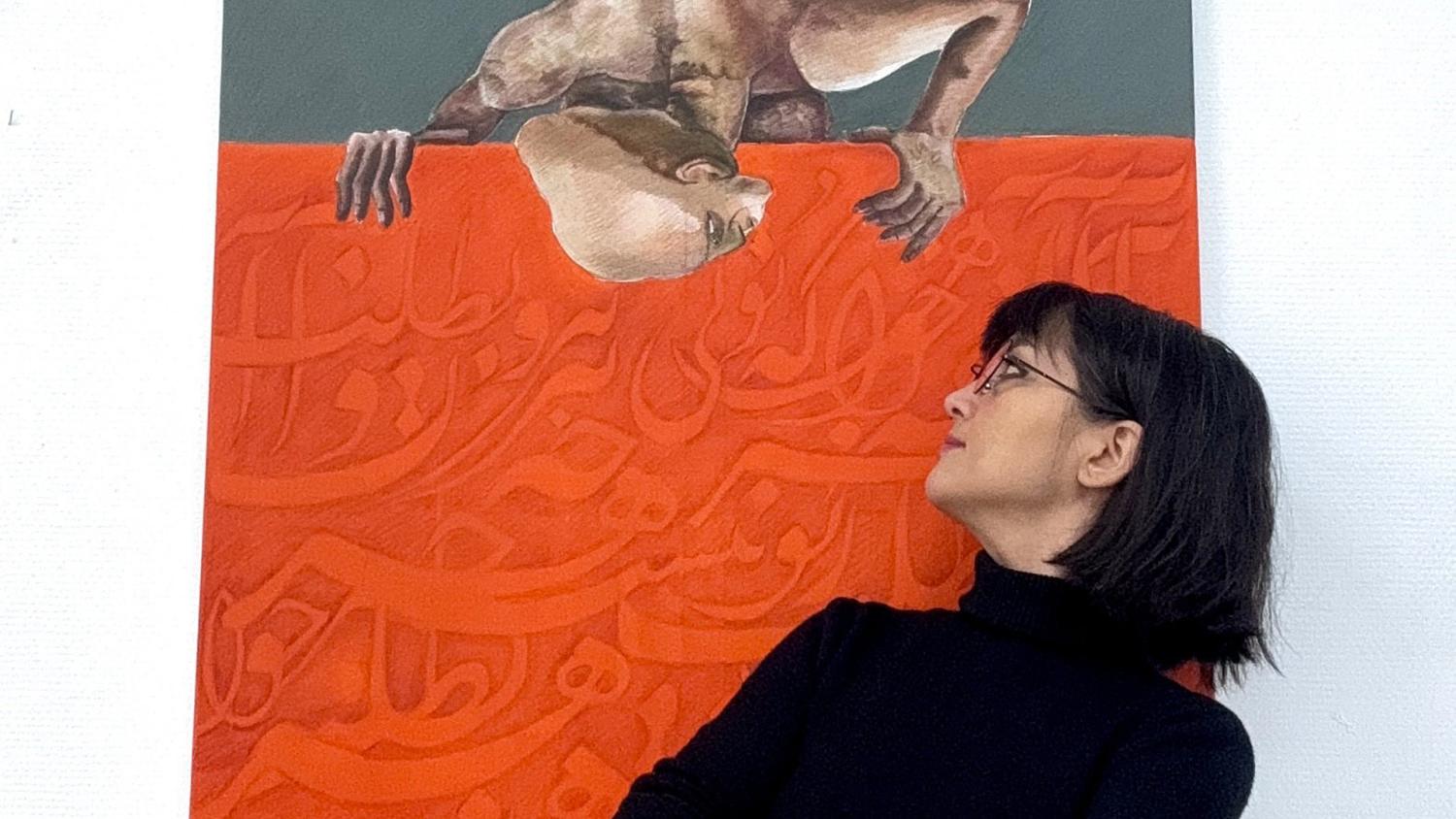 En kvinna i svart topp och glasögon tittar uppåt på en målning på väggen. Konstverket föreställer en person upp och ner ovanför arabisk skrift på röd bakgrund.