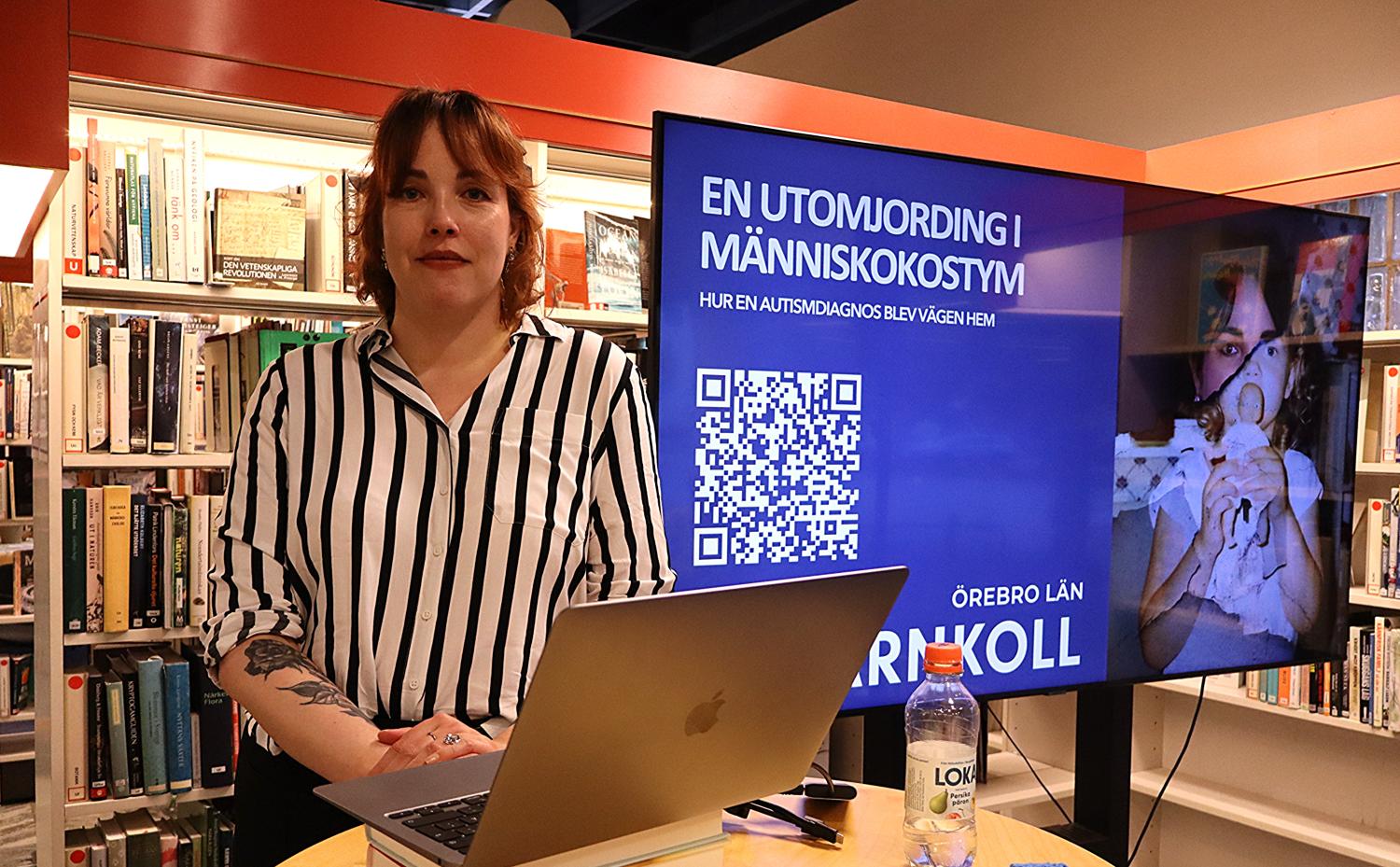 En kvinna står bredvid en bärbar dator och en presentationsskärm i ett bibliotek. På skärmen visas svensk text, en QR-kod och en bild av en person i kostym.