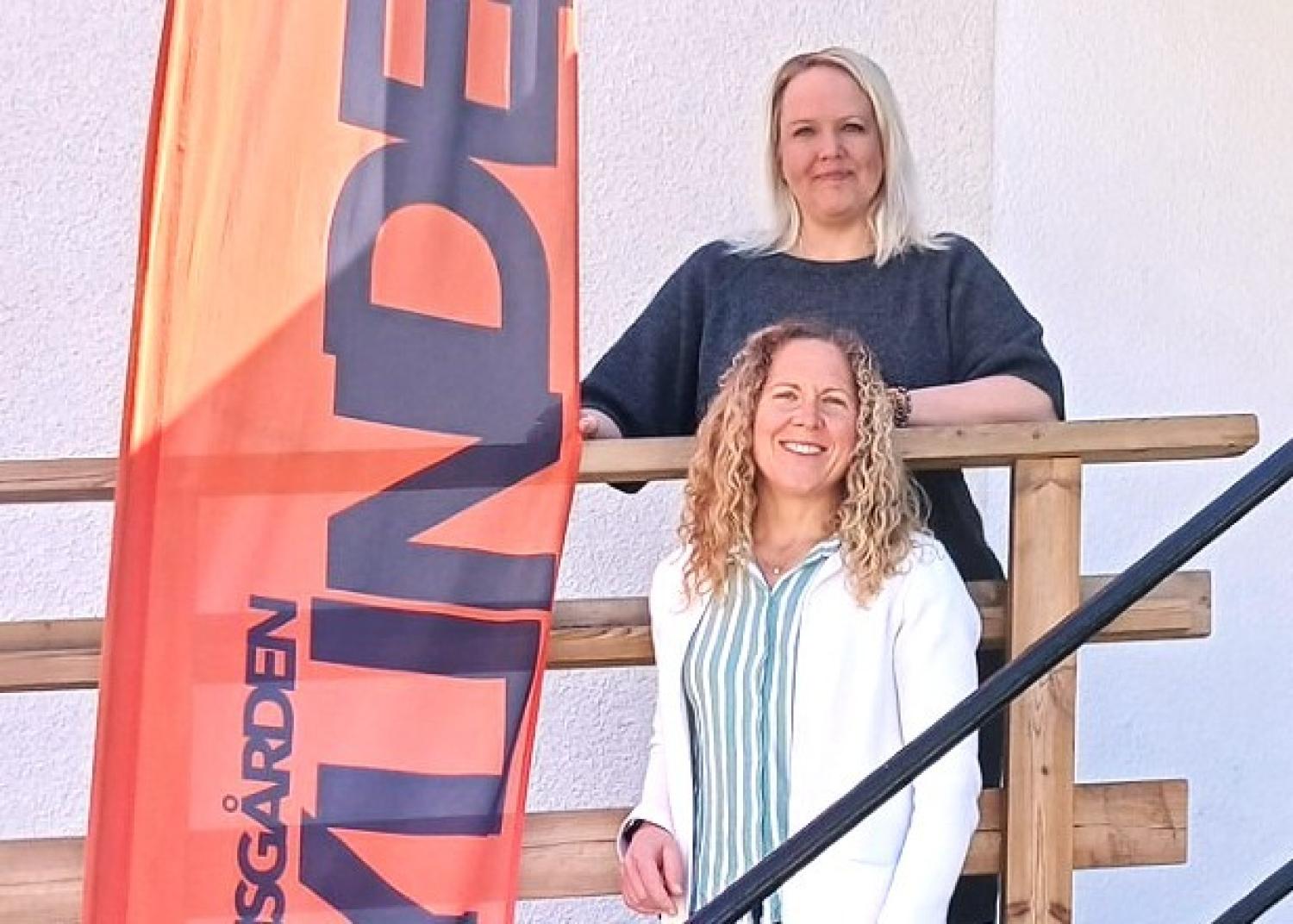 Två kvinnor står på en trätrappa bredvid en orange vertikal banner med svart text mot en vit vägg.