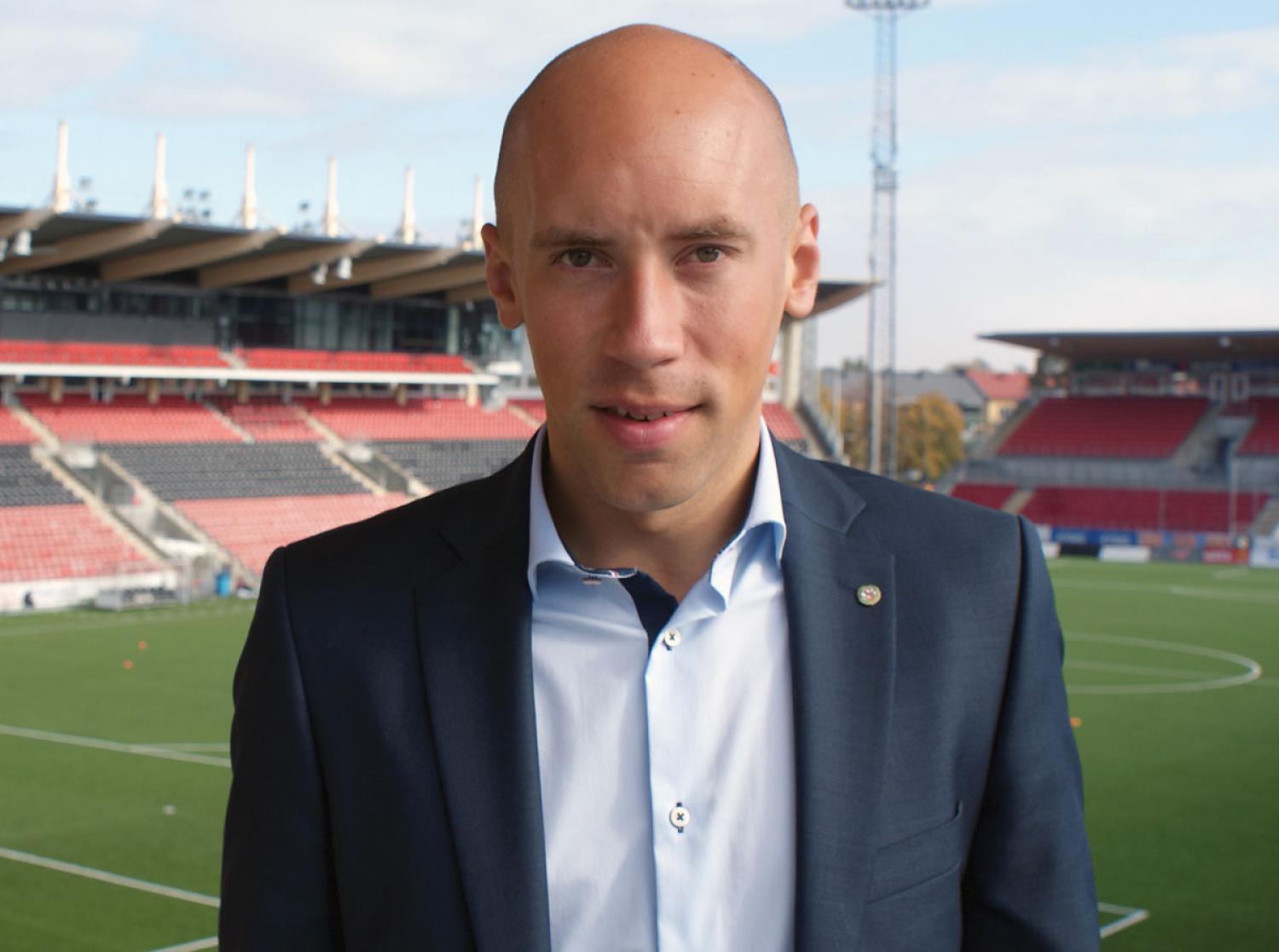 En person i kostym står framför en fotbollsplan med tomma stadionplatser.