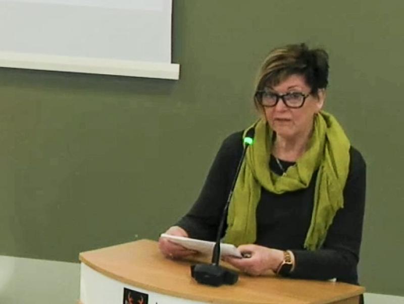 En kvinna med glasögon och grön halsduk talar på ett podium med mikrofon och håller papper i händerna.