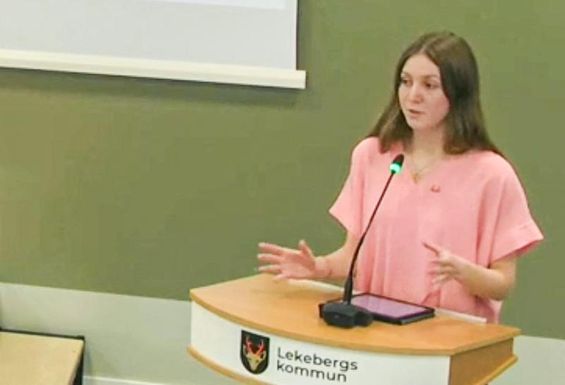 En person i rosa skjorta talar vid ett podium med mikrofon, märkt 