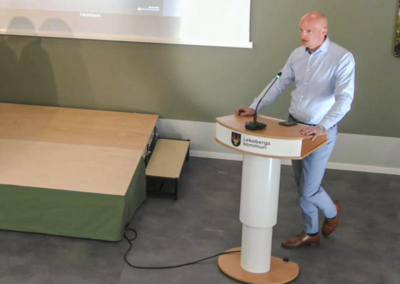 En man i ljusblå skjorta står vid ett podium med texten 
