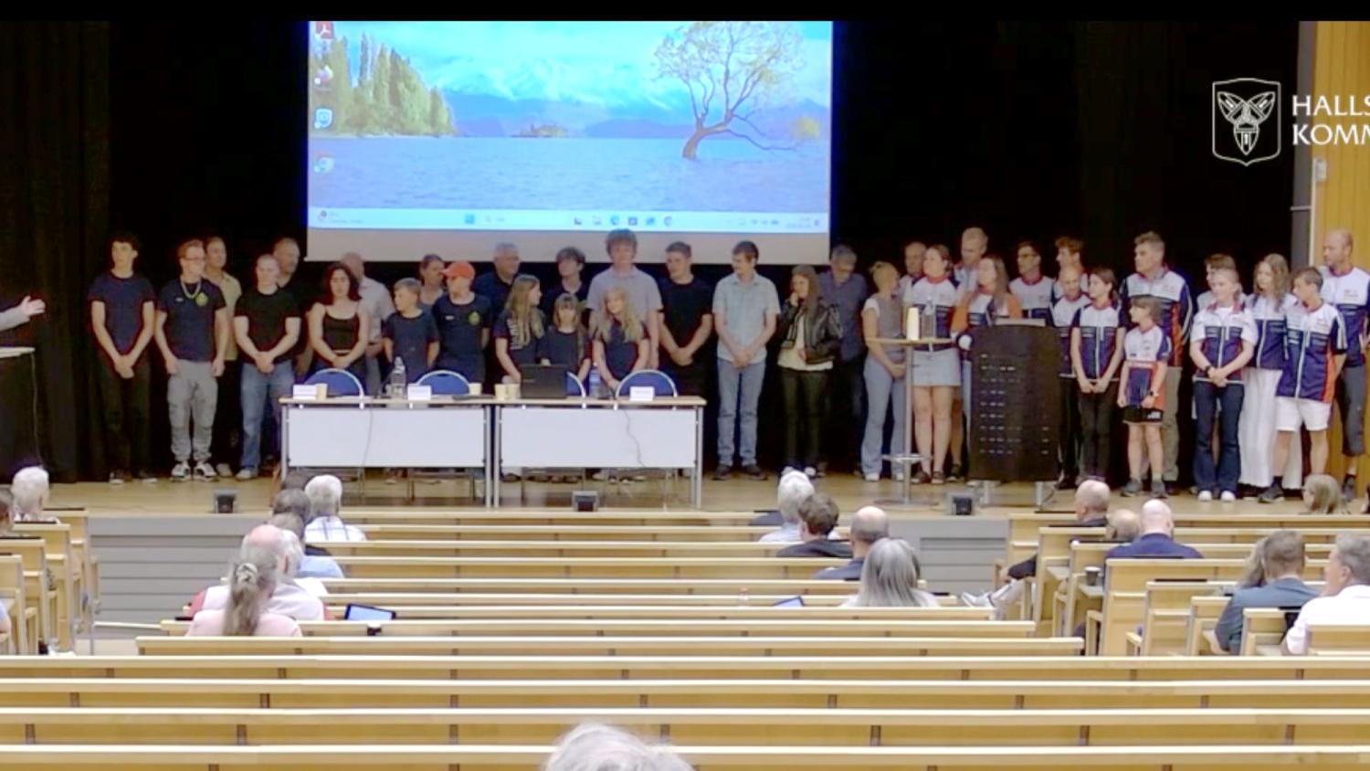 En grupp människor står på scenen framför en projektorduk och publik i en föreläsningssal med träbänkar.