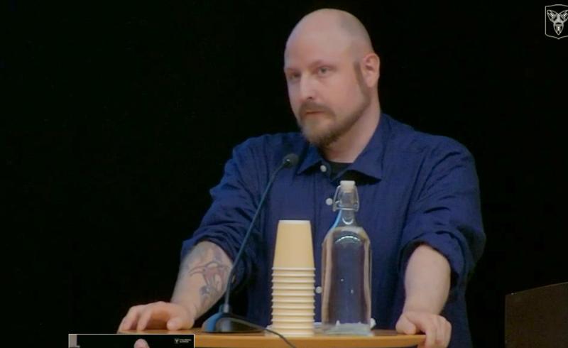 En person med tatuerad arm står på ett podium med staplade muggar och en glasflaska och talar i mikrofon mot en mörk bakgrund.