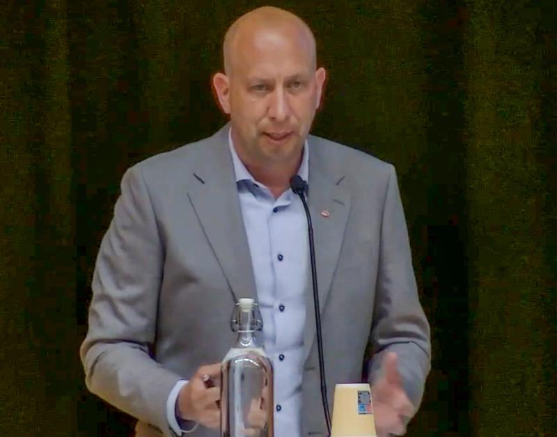 En man i grå kostym talar vid ett podium med en mikrofon, med en glasflaska och en engångsmugg framför sig mot en mörk bakgrund.