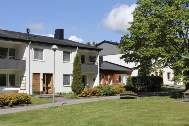 Tvåvåningshus med balkonger, omgivna av gröna gräsmattor, buskar, ett stort träd och en lyktstolpe under en delvis molnig himmel.