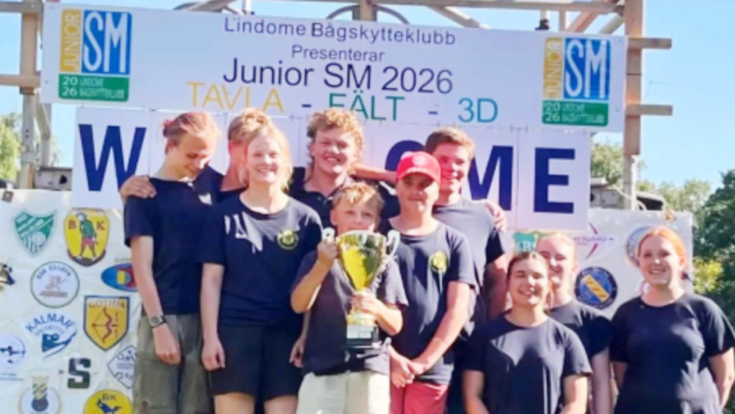 En grupp unga bågskyttar i likadana tröjor poserar utomhus och håller upp en pokal framför en välkomstskylt med svensk text om en bågskyttetävling för juniorer.