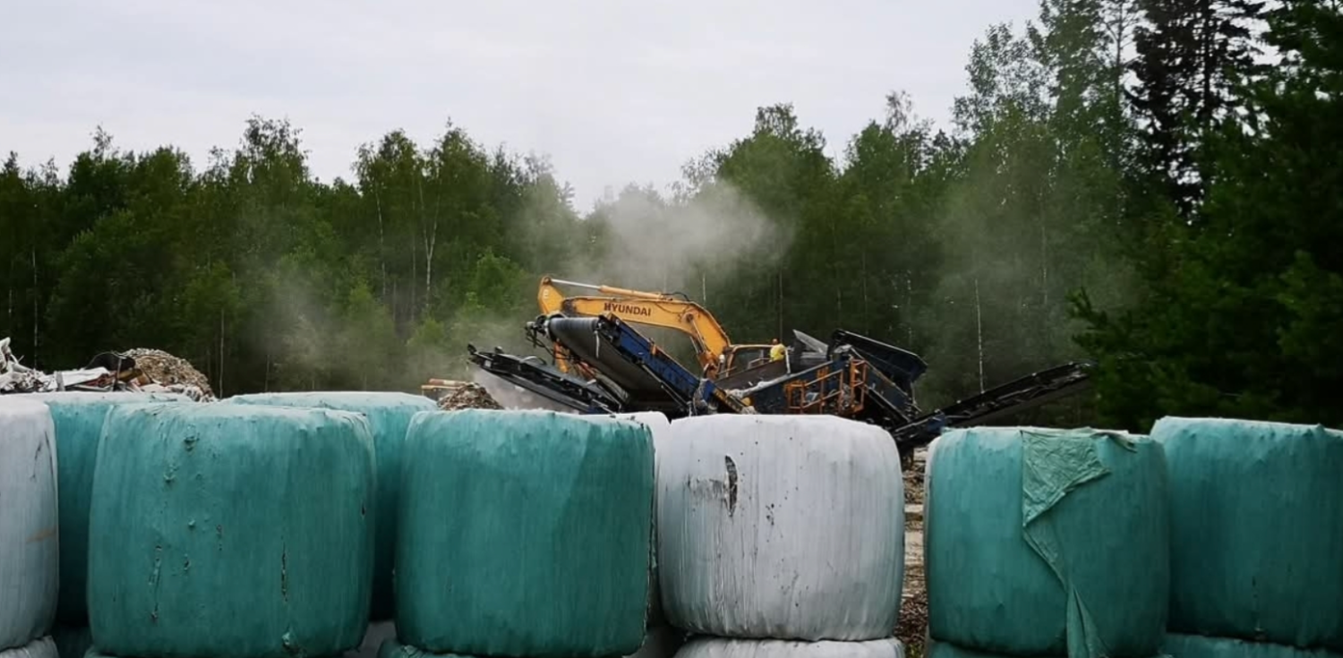 En grävmaskin arbetar på en bygg- eller avfallsplats och orsakar damm. I förgrunden står stora gröna och vita balar, inslagna i plast, staplade på varandra. I bakgrunden syns täta träd.