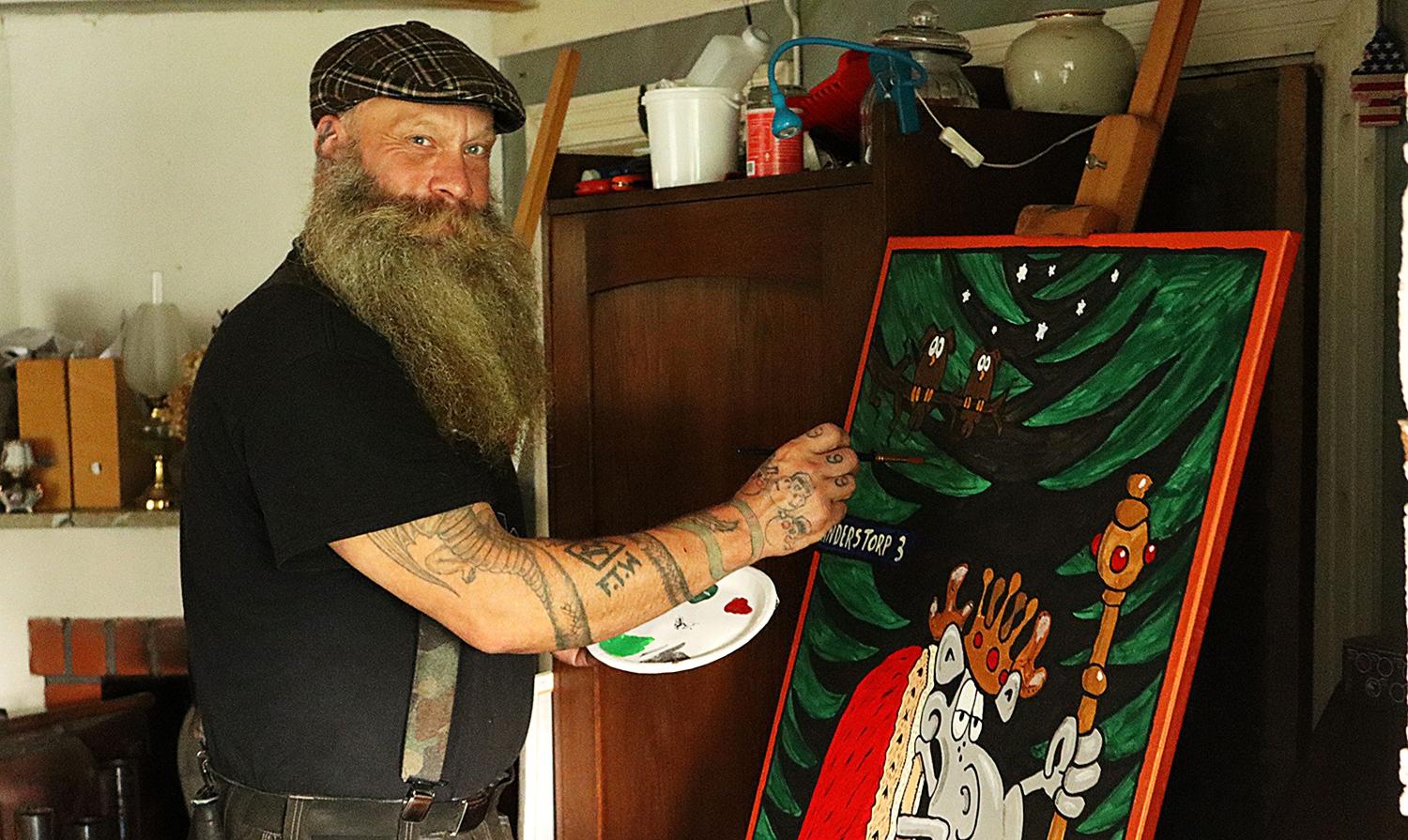En skäggig man med tatueringar målar ett färgstarkt konstverk i tecknad stil på en duk i ett rum fyllt med hushållsartiklar.