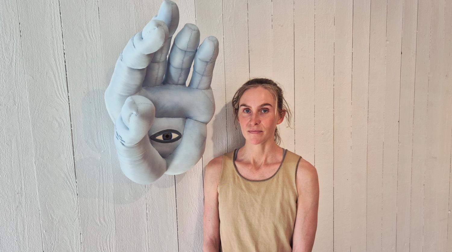 En kvinna i ett solbrunt linne står bredvid ett väggmonterat konstverk som föreställer en stor hand med ett öga i mitten av handflatan.