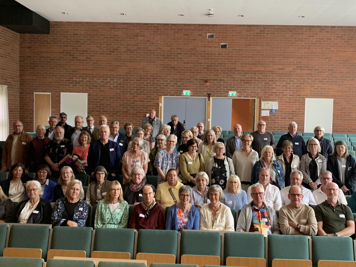 En stor grupp människor i olika åldrar poserar för ett gruppfoto i en aula med gröna sittplatser och tegelväggar.