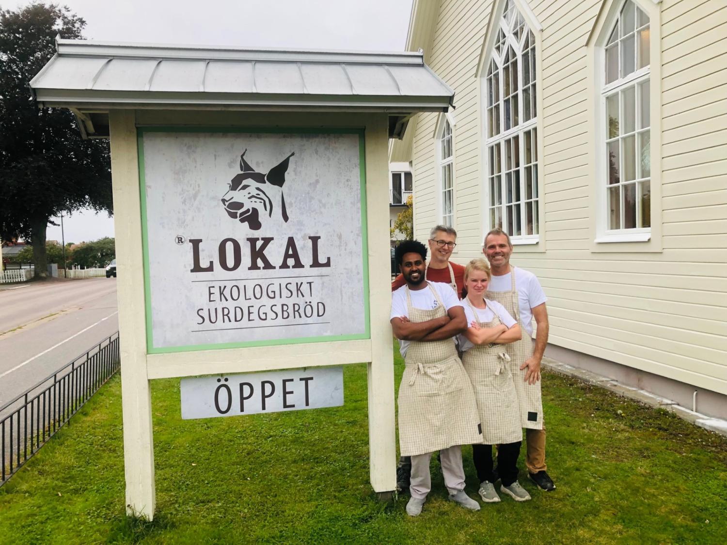 Fyra personer med förkläden står leende framför en skylt för 